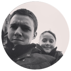 Black and white circular photo showing a man with a slightly furrowed brow and a young smiling child behind him. Both are wearing jackets and appear to be outdoors. The image is casual and candid in nature.